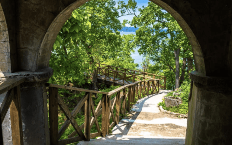 a path with wooden rails leading from a stone arch to a picturesque water body